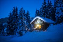 Áustria, Altenmarkt-Zauchensee, trenós, boneco de neve e árvore de Natal em casa de madeira iluminada na neve à noite — Fotografia de Stock