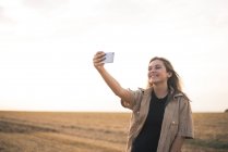 Молодая женщина стоит в поле, делает селфи на смартфоне — стоковое фото