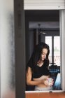 Jeune femme sérieuse avec une tasse de café à la fenêtre à la maison — Photo de stock