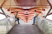 Молода гей-пара стоїть на сучасному пішохідному мосту, дивлячись один на одного — стокове фото