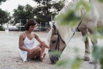 Femme souriante assise sur le sol près du cheval — Photo de stock