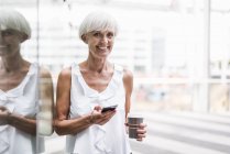 Портрет улыбающейся пожилой женщины с сотовым телефоном и кофе на открытом воздухе — стоковое фото