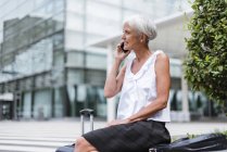 Mulher sênior sorridente com bagagem no telefone celular na cidade — Fotografia de Stock