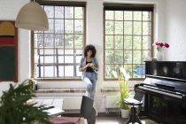 Frau steht am Fenster ihrer Dachgeschosswohnung und trinkt Kaffee — Stockfoto