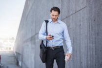 Uomo d'affari con auricolari a piedi lungo muro di cemento guardando smartphone — Foto stock