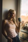 Jeune femme blonde tenant une tasse de café à la fenêtre — Photo de stock