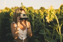 Mujer joven en un campo de girasoles tomando fotos con una cámara instantánea - foto de stock