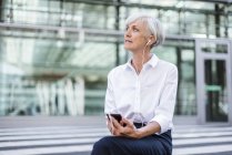 Femme d'affaires âgée assise devant une façade en verre avec smartphone et écouteurs — Photo de stock