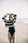 Donna bionda sulla spiaggia e posa sulla macchina fotografica, indossa maglione con strisce e costume da bagno nero con le mani dietro la testa — Foto stock
