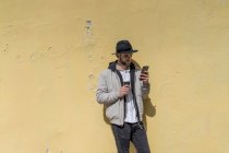 Бородатый мужчина в черной шляпе, опираясь на стену, используя смартфон, кофе, чтобы пойти — стоковое фото
