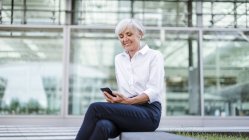 Empresária sênior sentada ao ar livre com smartphone e fones de ouvido — Fotografia de Stock