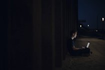 Mid adult man sitting cross-legged on ground, using laptop at night — Stock Photo