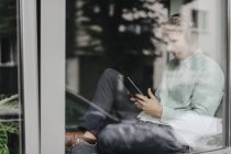 Jeune homme assis à la fenêtre, écoutant de la musique avec écouteurs et tablette numérique — Photo de stock