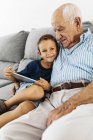 Retrato de niño feliz con tableta digital sentado con el abuelo en el sofá en casa - foto de stock