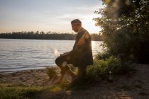 Empresário maduro sentado na margem do lago ao pôr-do-sol e trabalhando no laptop — Fotografia de Stock