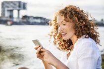 Alemanha, Colônia, retrato de uma jovem mulher assombrada sentada à beira do rio e olhando para o telefone celular — Fotografia de Stock