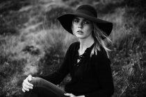 Retrato de jovem mulher vestindo chapéu preto e sentado ao ar livre e posando na câmera — Fotografia de Stock