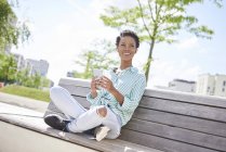 Portrait of smiling young woman with cell phone and earphones sitting on bench — Stock Photo