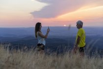 Padre mayor y la hija adulta tomando una foto del teléfono celular en la cima de la colina durante el atardecer - foto de stock