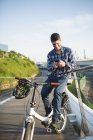 Smiling young man with bicycle using smartphone on bridge — Stock Photo