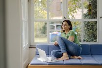 Lächelnde reife Frau sitzt zu Hause mit Laptop auf der Couch — Stockfoto