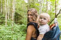 Porträt einer lächelnden Mutter, die mit dem kleinen Jungen im Rucksack im Wald wandert — Stockfoto
