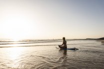 Junge Frau praktiziert Yoga am Strand, sitzt auf Surfbrett, meditiert — Stockfoto