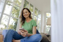 Усміхнена зріла жінка, сидячи на дивані вдома за допомогою мобільного телефону — стокове фото