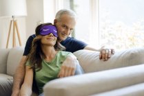 Smiling mature couple sitting on couch at home with woman wearing eye mask — Stock Photo