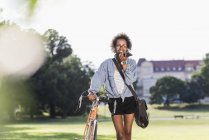Smiling young woman with cell phone and bicycle in park — Stock Photo