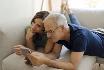 Sorridente coppia matura sdraiata sul divano a casa tablet condivisione — Foto stock