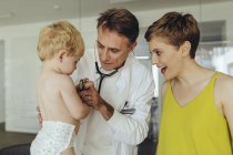 Pediatra examinando a un niño pequeño con estetoscopio, madre de pie junto a ellos - foto de stock