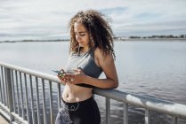 Giovane donna atletica indossa auricolari e utilizzando smartphone a riva del fiume — Foto stock