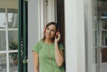 Mulher madura em pé na porta do terraço ouvindo música com fones de ouvido — Fotografia de Stock