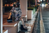 Homem do espaço sentado no banco no ponto de ônibus à noite e segurando o telefone celular — Fotografia de Stock