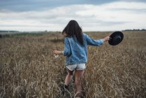 Rückansicht einer jungen Frau mit Getränk und Hut, die im Maisfeld spaziert — Stockfoto