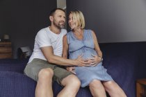 Mature man and pregnant mature wife sitting on bed and touching belly — Stock Photo