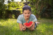 Porträt eines Mädchens, das auf einer Wiese im Garten liegt und Wassermelone isst — Stockfoto