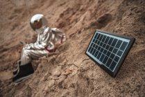 Spaceman sitting on nameless planet, charging device with solar panel — Stock Photo