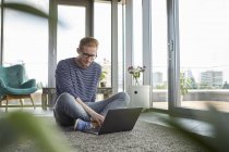 Молодой человек сидит дома на ковре и пользуется ноутбуком — стоковое фото