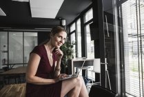 Femme d'affaires souriante au bureau portant une robe bordeaux à l'aide d'un ordinateur portable — Photo de stock