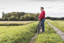 Homem idoso com bicicleta contemplando o caminho na paisagem rural — Fotografia de Stock