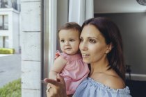 Lächelnde Mutter schaut mit kleiner Tochter zu Hause aus dem Fenster — Stockfoto