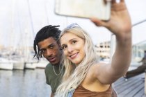 Portrait of multicultural young couple taking selfie with smartphone at harbour — Stock Photo