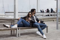 España, Barcelona, feliz pareja joven con teléfono celular descansando en un banco de la ciudad - foto de stock