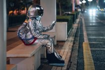 Homem do espaço sentado no banco no ponto de ônibus à noite e segurando o telefone celular — Fotografia de Stock