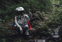 Spaceman crouching at brook in forest and holding rock — Stock Photo