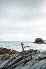 Junge Frau trägt Surfbrett an felsigem Strand auf See — Stockfoto