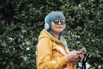 Retrato de mujer joven con el pelo azul teñido escuchando música con smartphone y auriculares - foto de stock