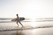 Jeune homme courant sur la plage, portant une planche de surf — Photo de stock